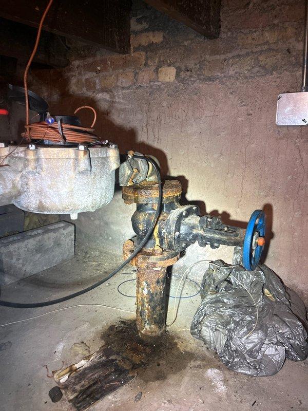 Completed assessment of aging plumbing infrastructure in basement utility area, identifying severely corroded vertical piping, rusted valve assembly with blue wheel handle, and deteriorating gas meter connections with brass and steel fittings. Documented existing conditions showing extensive oxidation on metal components, compromised pipe integrity, and outdated plumbing configuration requiring replacement to ensure safe operation and prevent potential leaks or system failure.