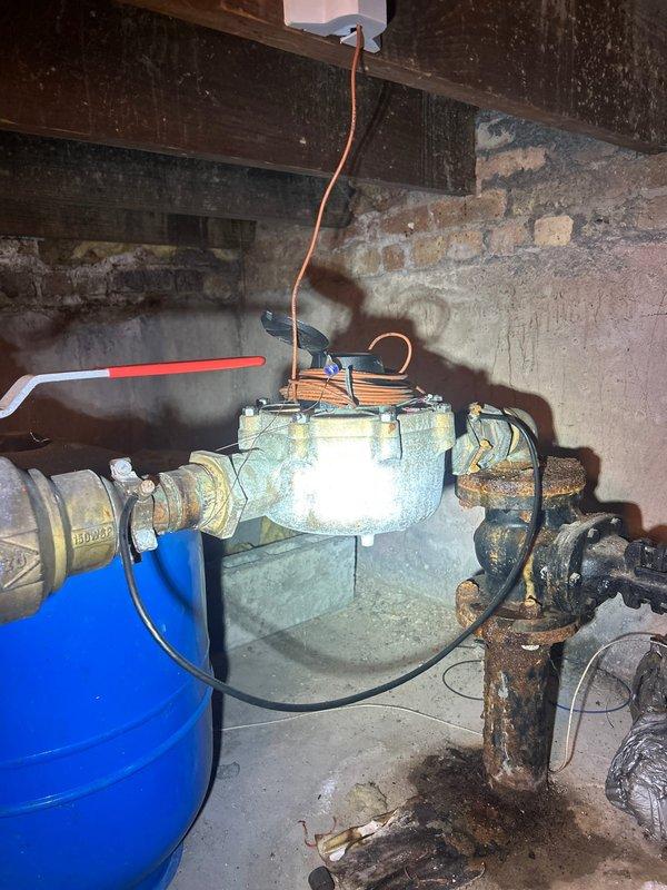 Completed assessment of aging plumbing infrastructure in basement utility area, identifying severely corroded vertical piping, rusted valve assembly with blue wheel handle, and deteriorating gas meter connections with brass and steel fittings. Documented existing conditions showing extensive oxidation on metal components, compromised pipe integrity, and outdated plumbing configuration requiring replacement to ensure safe operation and prevent potential leaks or system failure.