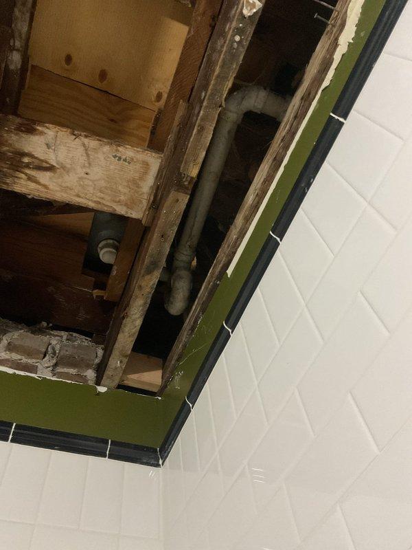 Responded to service call for ceiling water damage in bathroom. Upon inspection, discovered significant paint peeling on ceiling caused by active plumbing leak from above. Opened ceiling access point to expose source of water intrusion. Located compromised supply line running through wall cavity between floor joists. Removed section of ceiling material and exposed framing to access piping. Repaired leaking pipe connection and secured all plumbing fixtures. Documented existing conditions showing water-damaged ceiling paint, exposed wall framing, and piping configuration for insurance and restoration purposes.