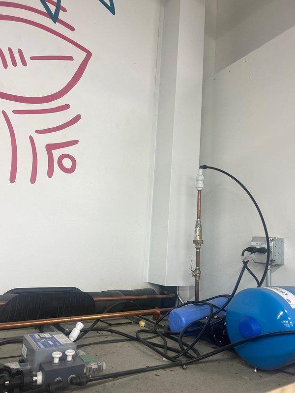 Completed installation of valve on water supply line and connected drainage system for new plumbing fixture. Installed blue expansion tank with copper piping connections, routed black flexible drain tubing, and integrated control unit with digital display panel for system monitoring and operation.