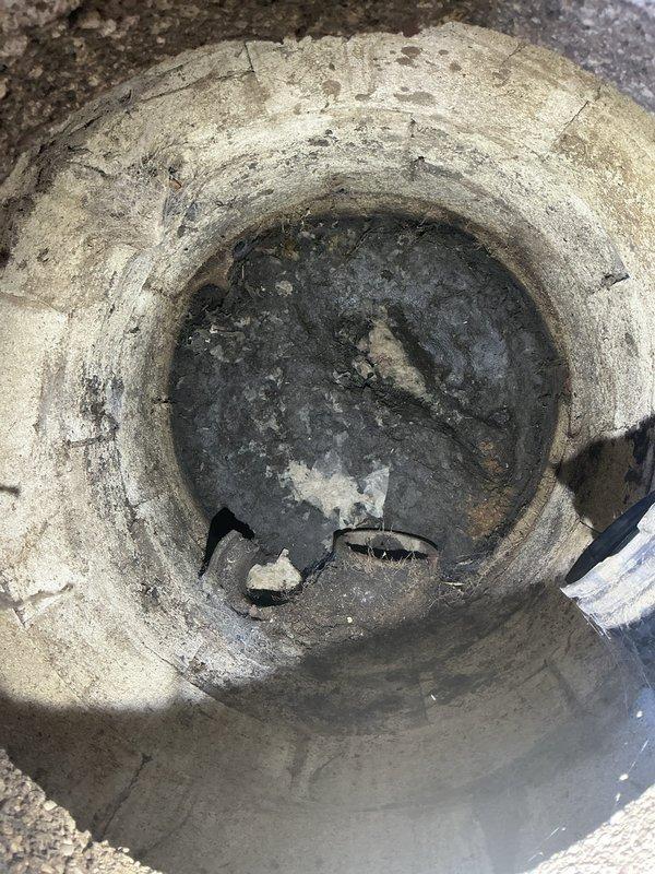 Inspected outdoor drainage system along building perimeter where concrete surface meets chain-link fence line. Assessed condition of floor drain with radial spoke pattern and investigated rusty pipe fitting embedded near fence base. Documented heavy sediment accumulation and standing water in circular concrete drainage pipe, with dark residue coating interior walls and debris collected at bottom. Photographed deteriorated conditions including surface weathering, dirt buildup at concrete-fence junction, and compromised drainage infrastructure requiring attention.