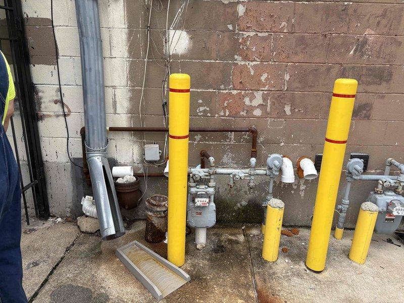 Conducted comprehensive site assessment of exterior gas meter installation and associated piping infrastructure for estimate preparation. Documented existing conditions including gas meter assembly, brown gas piping runs, multiple shut-off valves, and protective bollard system consisting of yellow cylindrical posts with red banding positioned to safeguard utility equipment from potential vehicle impact. Photographed deteriorating brick wall conditions, vertical gray conduit installation, exposed electrical wiring, mounted utility boxes, and concrete ground surface showing significant weathering and structural wear requiring consideration for future piping modifications or repairs.