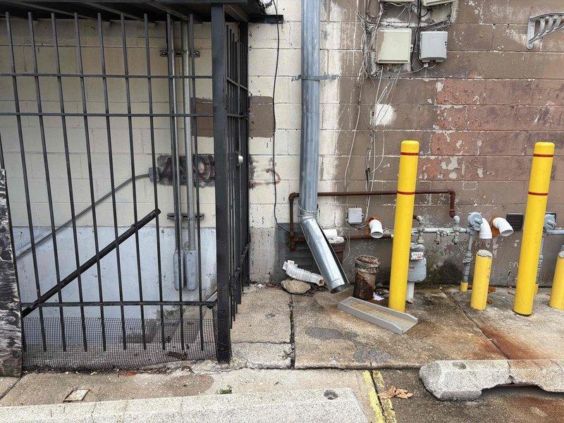 Conducted comprehensive site assessment of exterior gas meter installation and associated piping infrastructure for estimate preparation. Documented existing conditions including gas meter assembly, brown gas piping runs, multiple shut-off valves, and protective bollard system consisting of yellow cylindrical posts with red banding positioned to safeguard utility equipment from potential vehicle impact. Photographed deteriorating brick wall conditions, vertical gray conduit installation, exposed electrical wiring, mounted utility boxes, and concrete ground surface showing significant weathering and structural wear requiring consideration for future piping modifications or repairs.
