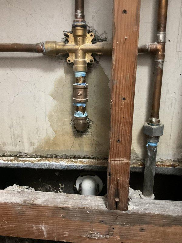 Responded to active water leak dripping into basement ceiling in 85-year-old residence. Upon inspection, identified deteriorated plumbing connections at brass cross-valve manifold with aged copper piping showing significant corrosion and tarnishing. Water staining on surrounding wall indicated ongoing moisture intrusion from compromised fittings. Accessed plumbing system through cabinet area beneath structure. Replaced failed connections using PVC pipe with proper primer application and secured flexible coupling with metal hose clamp to ensure watertight seal. Repaired vertical chrome pipe section with new blue plastic connectors at joints. Addressed single affected area and verified all connections for proper flow and eliminated active leak. System tested and confirmed operational with no further water intrusion detected.