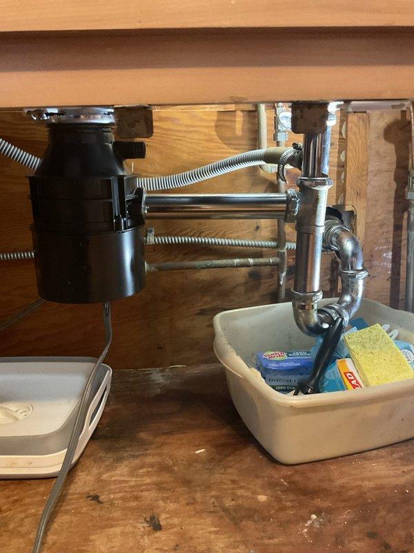 Responded to service call for garbage disposal leaking from bottom in 119-year-old residence. Upon inspection, identified active leak originating from lower housing seal of black garbage disposal unit mounted beneath kitchen sink. Documented existing water staining and moisture damage on cabinet floor and interior wood surfaces, indicating leak had been present for extended period. Assessed disposal unit mounting hardware, drain connections, and flexible corrugated hoses for integrity. Due to age and location of leak at bottom seal, determined disposal unit required replacement as bottom seal failures are not repairable. Photographed existing installation configuration showing disposal unit, chrome plumbing connections, flexible drain hoses, and cabinet interior condition for documentation purposes. Provided customer with assessment findings and replacement recommendations to resolve leak issue and prevent further cabinet damage.