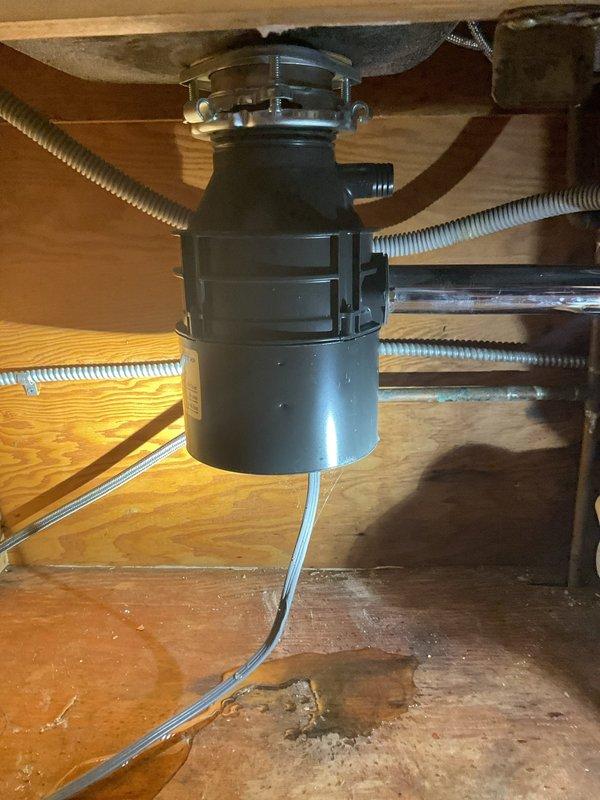 Responded to service call for garbage disposal leaking from bottom in 119-year-old residence. Upon inspection, identified active leak originating from lower housing seal of black garbage disposal unit mounted beneath kitchen sink. Documented existing water staining and moisture damage on cabinet floor and interior wood surfaces, indicating leak had been present for extended period. Assessed disposal unit mounting hardware, drain connections, and flexible corrugated hoses for integrity. Due to age and location of leak at bottom seal, determined disposal unit required replacement as bottom seal failures are not repairable. Photographed existing installation configuration showing disposal unit, chrome plumbing connections, flexible drain hoses, and cabinet interior condition for documentation purposes. Provided customer with assessment findings and replacement recommendations to resolve leak issue and prevent further cabinet damage.