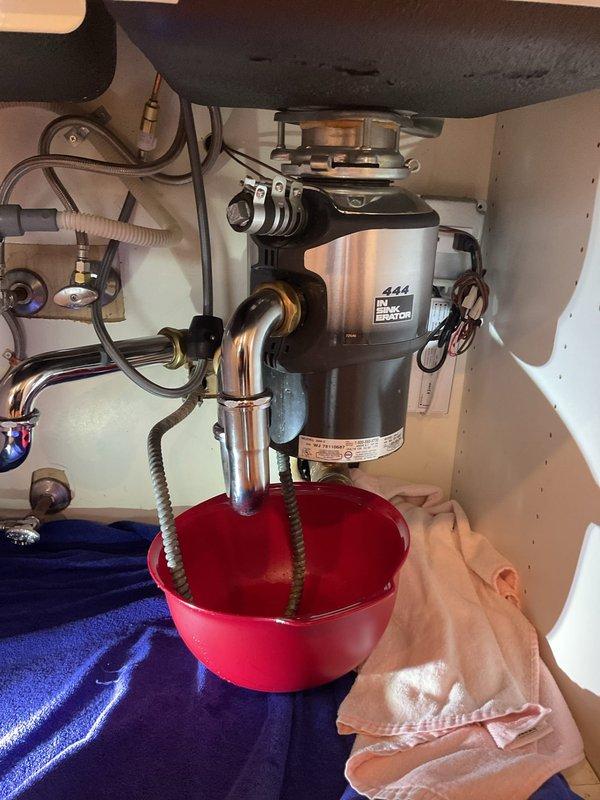 Responded to water leak complaint originating from kitchen sink area affecting unit below. Upon arrival, performed visual inspection of under-sink plumbing system including garbage disposal unit, drain connections, and supply lines. Identified active leak at sink drain assembly during diagnostic assessment. Observed InSinkErator disposal unit with associated P-trap configuration and supply line connections. Placed collection bucket beneath leak source to prevent further water damage to lower unit. Documented existing plumbing conditions with photographic evidence showing disposal mounting, drain pipe connections, and leak collection setup for customer records and potential follow-up repair authorization.