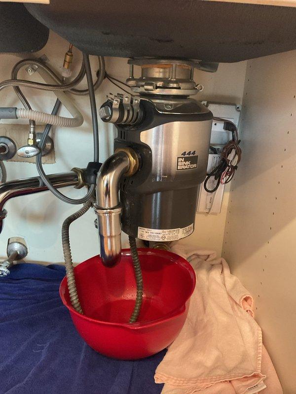 Responded to water leak complaint originating from kitchen sink area affecting unit below. Upon arrival, performed visual inspection of under-sink plumbing system including garbage disposal unit, drain connections, and supply lines. Identified active leak at sink drain assembly during diagnostic assessment. Observed InSinkErator disposal unit with associated P-trap configuration and supply line connections. Placed collection bucket beneath leak source to prevent further water damage to lower unit. Documented existing plumbing conditions with photographic evidence showing disposal mounting, drain pipe connections, and leak collection setup for customer records and potential follow-up repair authorization.