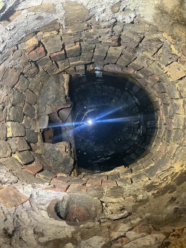 Responded to service call for removal of deteriorated brick-lined catch basin. Upon arrival, documented existing conditions showing aged circular brick structure with significant mortar deterioration and weathered masonry. The catch basin shaft exhibited traditional arched brickwork construction with loose and displaced bricks throughout. Basin contained standing water at bottom of shaft. Successfully removed old catch basin structure from property. Work area located in unfinished utility space with exposed wooden walls and concrete flooring. All debris from removal process cleared from site and surrounding area left clean.