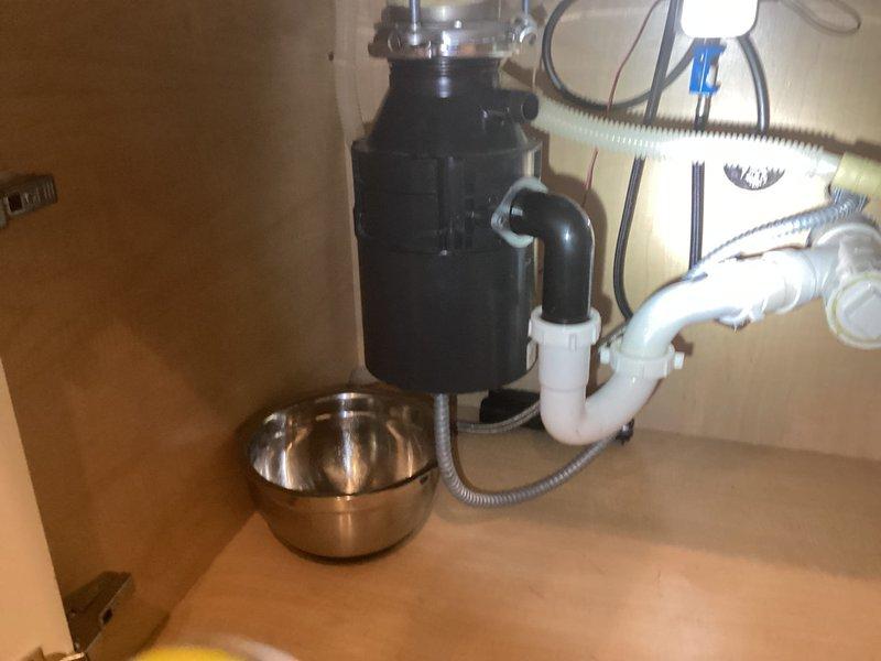 Responded to service call for garbage disposal leak at mounting screw location. Upon inspection, identified active water seepage from bottom of disposal unit at one of the mounting screws connecting the unit to the sink flange. Assessed the disposal unit, mounting assembly, and surrounding plumbing connections including the discharge elbow and P-trap configuration. Documented existing conditions showing black garbage disposal unit with metallic mounting flange, connected white PVC drainage system, and dishwasher drain line. Determined leak was originating from compromised seal at mounting bracket. Tightened mounting screws and verified integrity of upper mounting assembly and sink flange connection. Tested disposal operation and checked for proper drainage flow through discharge tube to P-trap. Confirmed leak resolution and proper function of all components.