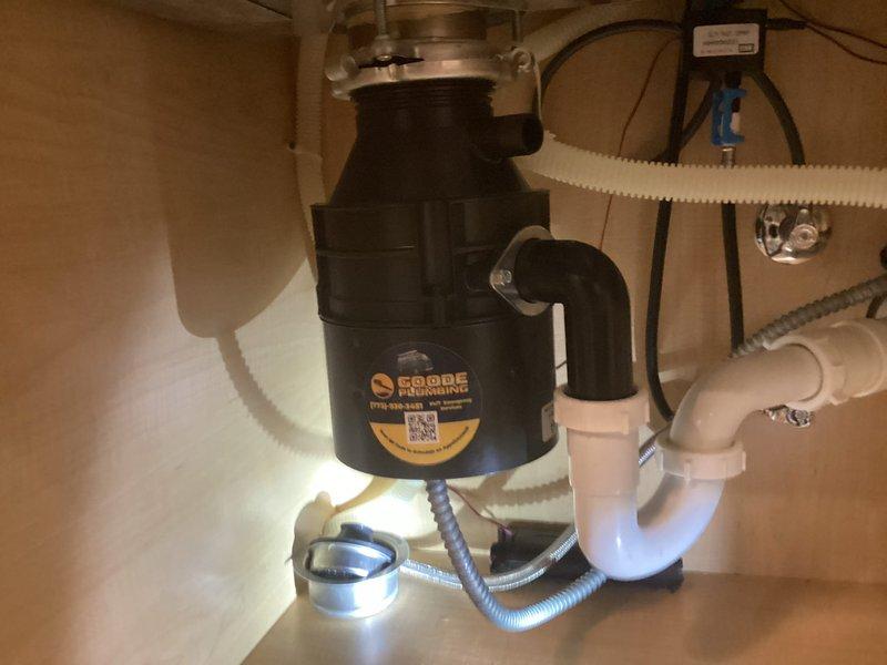 Responded to service call for garbage disposal leak at mounting screw location. Upon inspection, identified active water seepage from bottom of disposal unit at one of the mounting screws connecting the unit to the sink flange. Assessed the disposal unit, mounting assembly, and surrounding plumbing connections including the discharge elbow and P-trap configuration. Documented existing conditions showing black garbage disposal unit with metallic mounting flange, connected white PVC drainage system, and dishwasher drain line. Determined leak was originating from compromised seal at mounting bracket. Tightened mounting screws and verified integrity of upper mounting assembly and sink flange connection. Tested disposal operation and checked for proper drainage flow through discharge tube to P-trap. Confirmed leak resolution and proper function of all components.
