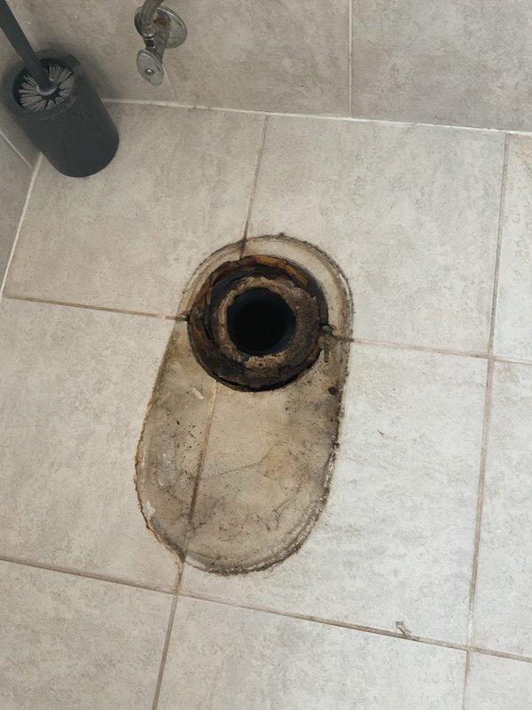 Responded to priority water leak on second floor toilet causing water intrusion into first floor bathroom ceiling in century-old home. Upon inspection, identified compromised toilet flange and wax ring seal as primary source of leakage. Evidence of chronic water seepage visible through significant staining and discoloration on bathroom floor tiles surrounding the exposed drain opening. Removed existing toilet to access deteriorated flange assembly. Documented existing conditions showing rust-colored water damage penetrating tile and grout around drain area, indicating prolonged leak activity. Assessed structural integrity of subflooring and underlying plumbing connections to determine extent of water damage and necessary repairs for proper toilet reinstallation and leak remediation.