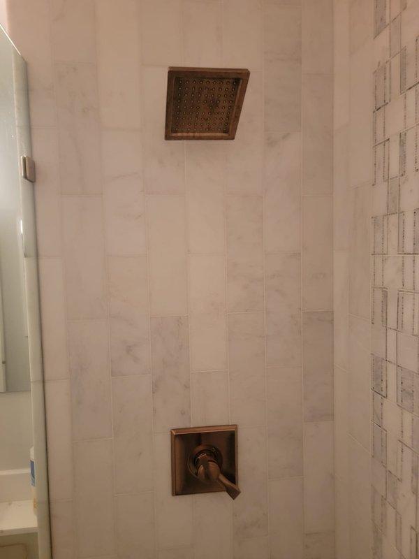 Completed installation of new shower fixtures in tiled shower enclosure. Installed oil-rubbed bronze rain shower head and matching shower valve control with triangular handle on square escutcheon plate. All fixtures properly mounted and sealed against light beige vertical tile on main wall and white marble-patterned tile surfaces, ensuring proper water flow and drainage functionality.