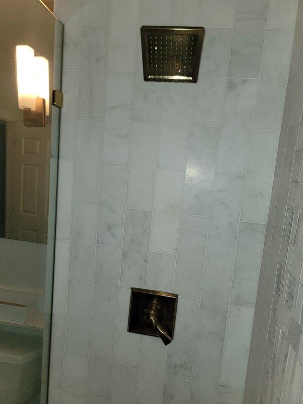 Completed installation of new shower fixtures in tiled shower enclosure. Installed oil-rubbed bronze rain shower head and matching shower valve control with triangular handle on square escutcheon plate. All fixtures properly mounted and sealed against light beige vertical tile on main wall and white marble-patterned tile surfaces, ensuring proper water flow and drainage functionality.