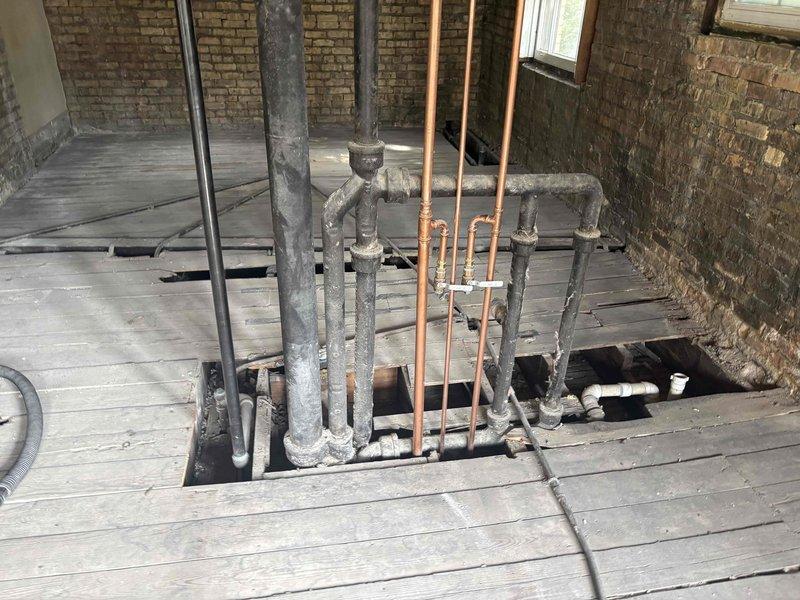 Completed comprehensive plumbing estimate for residential renovation project involving multiple drain line modifications and installations. Assessed requirements for repositioning shower drain, dividing existing soil stack to accommodate two toilet fixtures, adding dedicated drain line for washer and dryer hookup, and connecting various pipe sections throughout the work area. Documented existing plumbing infrastructure including aged galvanized pipes and newer copper piping emerging through concrete floor openings in basement area with exposed brick walls and deteriorated wooden deck structure requiring replacement.