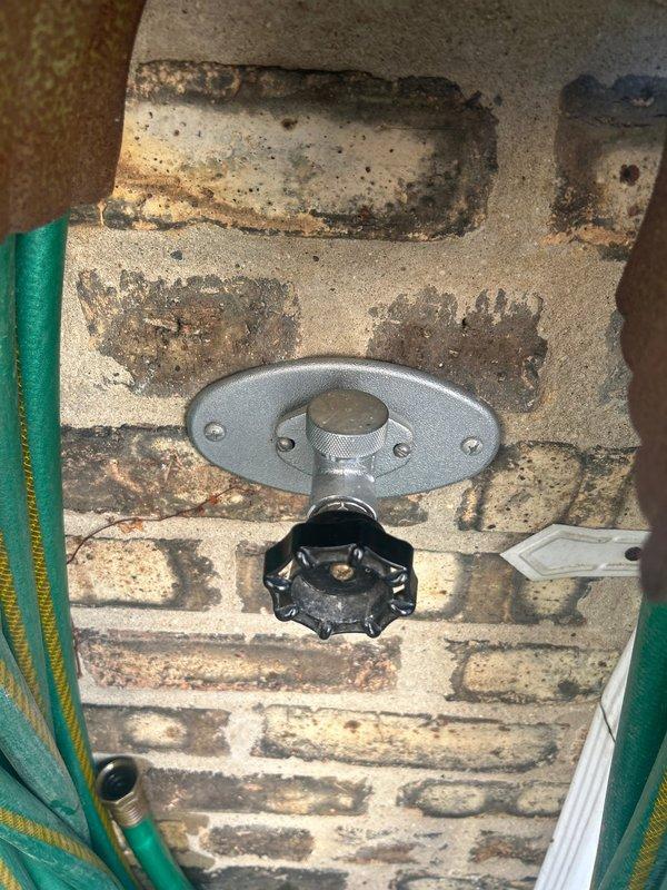 Responded to early morning service call for outdoor spigot inspection and assessment. Upon arrival, evaluated exterior-mounted hose bib installation on weathered brick wall showing significant mortar deterioration and moisture staining around fixture area. Documented existing conditions of galvanized valve assembly with oval mounting plate and operational control knob. Inspected mounting hardware security and verified proper fixture attachment to compromised masonry substrate. Photographed evidence of extensive brick and mortar degradation surrounding the installation point, noting dark staining patterns indicative of prolonged moisture exposure and potential water intrusion issues affecting wall integrity.