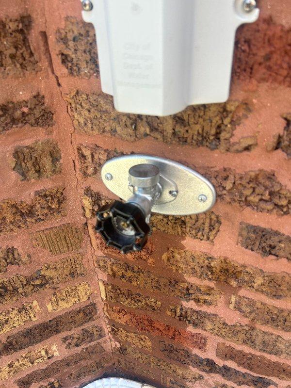 Responded to early morning service call for outdoor spigot inspection and assessment. Upon arrival, evaluated exterior-mounted hose bib installation on weathered brick wall showing significant mortar deterioration and moisture staining around fixture area. Documented existing conditions of galvanized valve assembly with oval mounting plate and operational control knob. Inspected mounting hardware security and verified proper fixture attachment to compromised masonry substrate. Photographed evidence of extensive brick and mortar degradation surrounding the installation point, noting dark staining patterns indicative of prolonged moisture exposure and potential water intrusion issues affecting wall integrity.