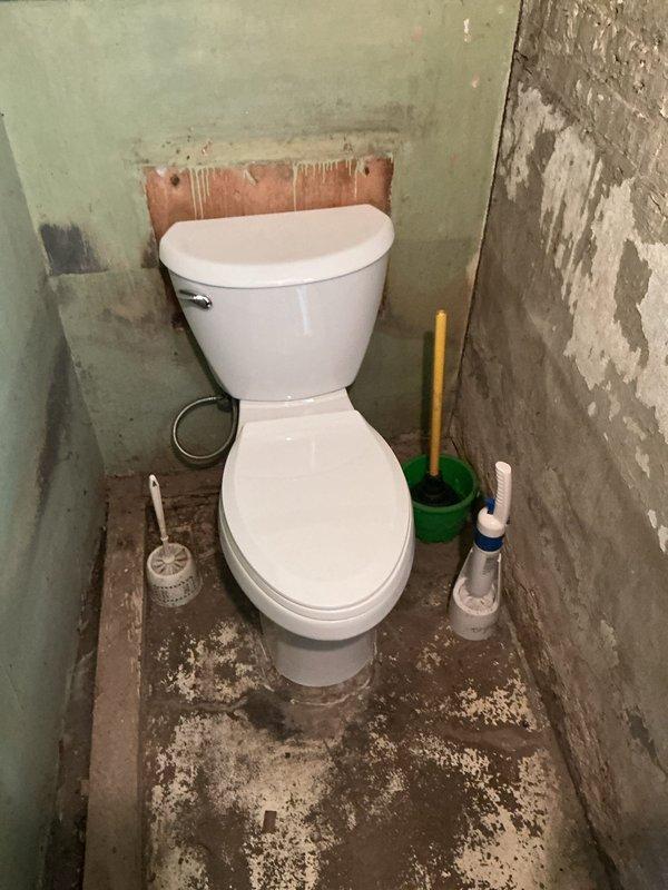 Removed existing toilet from basement bathroom and identified multiple code and structural deficiencies requiring permit work. Found cracked toilet flange with only two anchor bolts causing fixture instability and water seepage. Inspection revealed deteriorated clay drain pipe requiring replacement with PVC, necessitating concrete floor demolition and subsequent repair. Assessment determined new wax ring seal and proper flange installation will be required upon completion of rough-in work. Documented conditions with photographs showing corroded flange components, damaged flooring, and deteriorated wall surfaces for permit application and scope development.