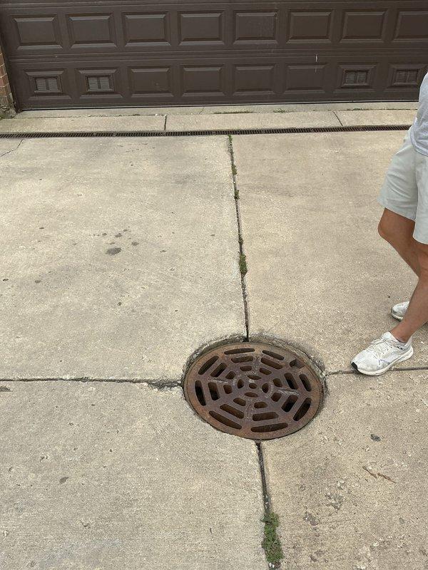 Inspected and assessed driveway drainage system including storm drain grate and surrounding concrete surfaces. Documented condition of metal drain cover with decorative pattern and evaluated concrete expansion joints showing vegetation growth and minor surface deterioration. Completed photographic documentation of drainage infrastructure and driveway condition for service records.