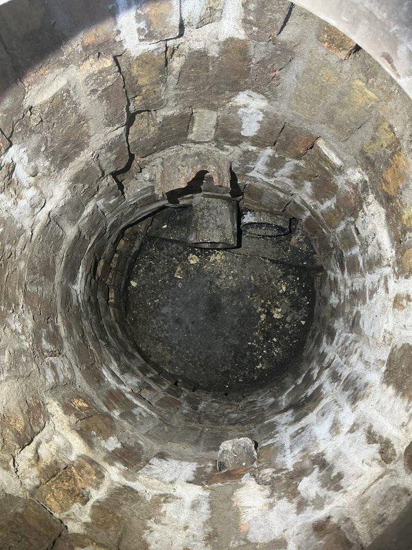 Responded to customer request for main drain cleanout installation and hydrojetting estimates. Inspected existing drainage access point located in outdoor utility structure with plywood construction and foam mat flooring. Evaluated circular floor access opening leading to deteriorated concrete sump pit approximately two feet in diameter. Documented extensive concrete degradation including vertical cracking, efflorescence, water staining, and mineral deposits throughout cylindrical structure. Observed standing water and sediment accumulation at bottom of pit with visible mounting hardware on interior walls. Assessed feasibility of installing new cleanout port at this exterior location versus alternative basement installation beneath concrete floor. Photographed existing conditions showing weathered concrete infrastructure and access limitations within wooden shed structure to provide accurate estimate for cleanout installation and hydrojetting services.