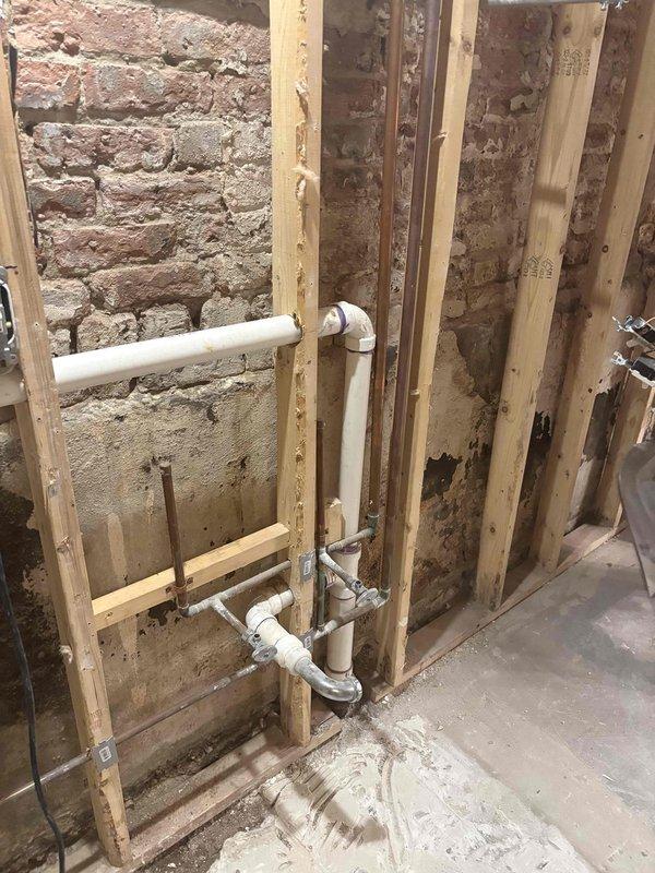Provided comprehensive plumbing estimate for remodel project involving drainage system installation in older building with exposed brick walls. Assessment included evaluation of existing floor drain, new PVC waste line routing through wall cavities, and integration with structural framing requirements. Documented current conditions of concrete flooring, drain pan placement, and vertical pipe runs to support detailed piping scope for renovation work.