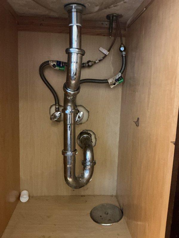 Responded to emergency service call after drain rodding equipment punctured neighboring unit's drainage pipe. Upon arrival, inspected under-sink plumbing assembly and confirmed damage to shared drain line. Documented existing conditions including chrome P-trap configuration, supply line connections, and cabinet interior. Photographed affected plumbing system showing standard residential drain assembly with J-trap, tailpiece, shut-off valves, and flexible supply hoses. Coordinated with property management regarding cross-unit pipe damage and repair requirements.