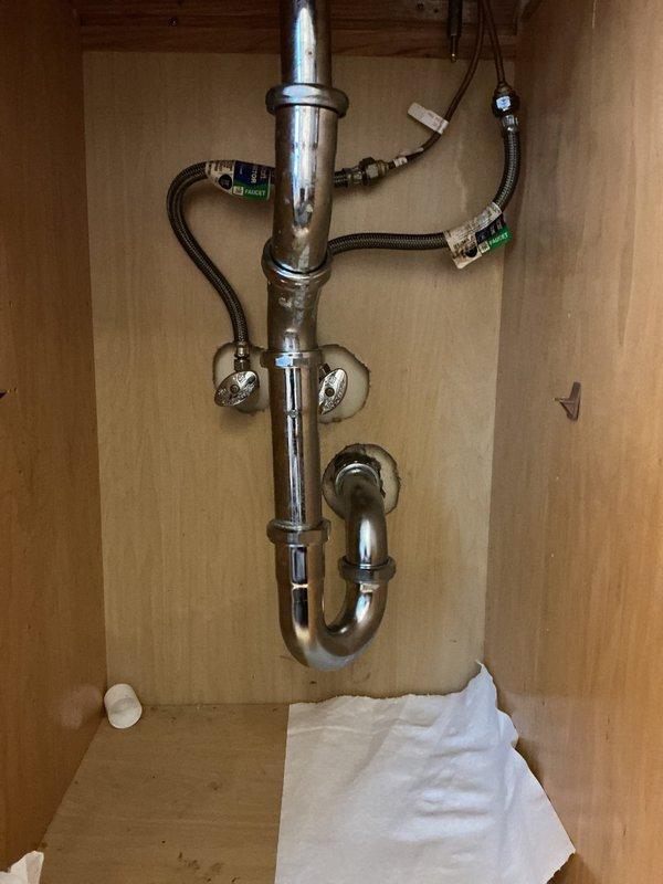 Responded to emergency service call after drain rodding equipment punctured neighboring unit's drainage pipe. Upon arrival, inspected under-sink plumbing assembly and confirmed damage to shared drain line. Documented existing conditions including chrome P-trap configuration, supply line connections, and cabinet interior. Photographed affected plumbing system showing standard residential drain assembly with J-trap, tailpiece, shut-off valves, and flexible supply hoses. Coordinated with property management regarding cross-unit pipe damage and repair requirements.