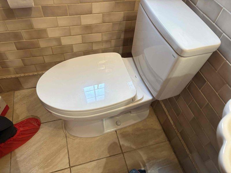 Completed installation of a new two-piece white porcelain toilet in residential bathroom with brown and beige subway tile walls and tan ceramic floor tiles. Toilet was properly positioned, secured, and tested to ensure proper function and no leaks upon completion.