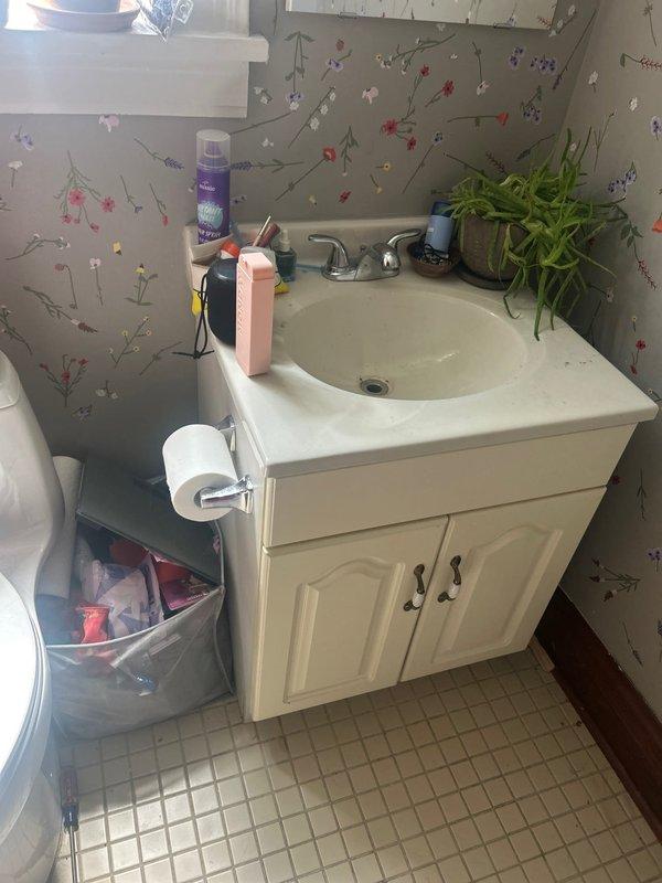 Conducted callback inspection of bathroom sink plumbing system to assess requirements for re-piping work. Documented existing vanity configuration including white cabinet with cream countertop, chrome faucet fixture, and current drain assembly. Examined under-sink plumbing components including white PVC drain pipes, chrome P-trap, dual chrome shut-off valves with flexible braided supply lines, and wall-mounted connections. Photographed visible cabinet deterioration and water staining on interior surfaces indicating previous moisture issues. Prepared documentation for follow-up estimate on complete re-piping of bathroom sink to restore full service functionality.