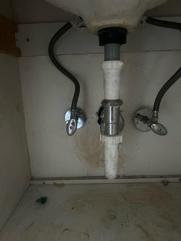 Conducted callback inspection of bathroom sink plumbing system to assess requirements for re-piping work. Documented existing vanity configuration including white cabinet with cream countertop, chrome faucet fixture, and current drain assembly. Examined under-sink plumbing components including white PVC drain pipes, chrome P-trap, dual chrome shut-off valves with flexible braided supply lines, and wall-mounted connections. Photographed visible cabinet deterioration and water staining on interior surfaces indicating previous moisture issues. Prepared documentation for follow-up estimate on complete re-piping of bathroom sink to restore full service functionality.