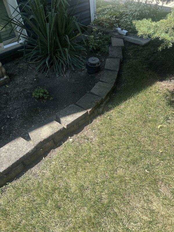 Conducted comprehensive site assessment for plumbing piping estimate at residential property. Documented exterior conditions including lawn irrigation system components and underground utility access points. Photographed existing landscape features, displaced concrete edging blocks along garden border, and circular metal access cover embedded in lawn area. Assessed accessibility for potential pipe routing and identified existing irrigation infrastructure including sprinkler heads in mulched garden beds. Evaluated ground conditions and documented current state of utility access points to determine scope of work for piping installation or repair project.