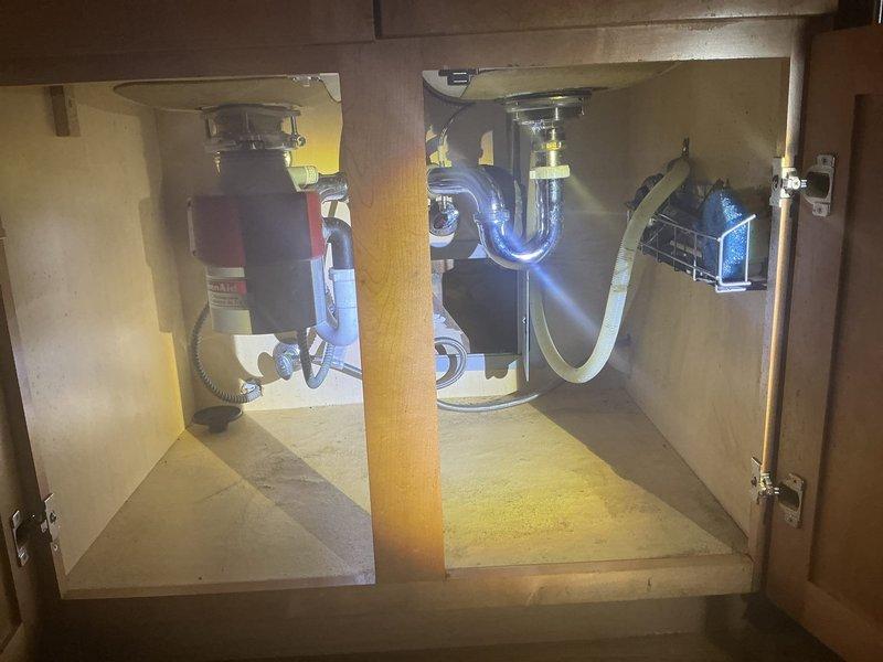 Conducted residential drain and sewer estimate for kitchen sink plumbing system. Assessed existing configuration including garbage disposal unit, P-trap assembly, and drainage piping beneath undermount sink installation. Documented current plumbing conditions and photographed cabinet interior showing disposal unit, PVC drainage connections, chrome P-trap, and dishwasher drain line routing for estimate preparation.