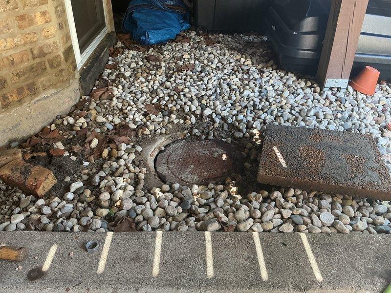 Accessed outdoor plumbing drain system located in side yard landscaping area. Removed and displaced decorative river rock ground cover to expose circular drain grate and surrounding infrastructure for inspection and repair assessment. Cleared work area including concrete pavers to provide full access to drainage components. Repair solution presented and approved by property owner for underground plumbing system servicing the residential structure.