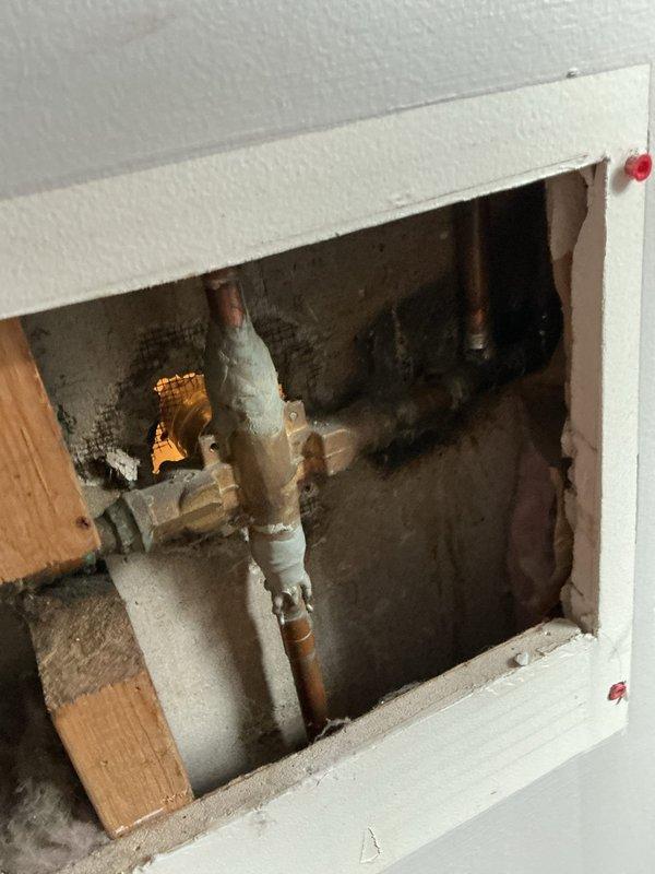 Responded to service call for shower valve issues. Upon inspection, found the shower stopper mechanism was loose and the valve handle trim piece had broken off, preventing proper operation of the fixture. Accessed the plumbing through existing wall access panel, exposing aged copper supply lines with visible oxidation and foam pipe insulation. Documented the condition of the valve assembly behind the white subway tile wall, noting the valve stem and internal components requiring attention. Photographed the exposed plumbing infrastructure and damaged valve hardware for estimate preparation and customer review.