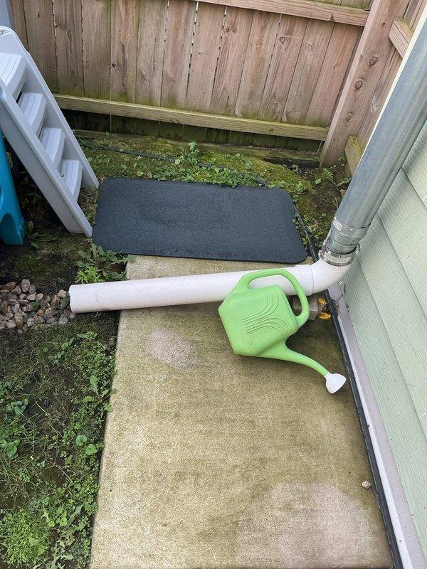 Conducted residential drain and sewer estimate for downspout drainage issue where the existing catch basin under the house had been closed off. The property currently has water directed from the downspout to a temporary solution using a green watering can connected to white PVC piping from a gray cylindrical drainage pipe mounted on the building exterior. Assessed the concrete walkway area showing weathering and drainage patterns, documented the current makeshift drainage setup, and evaluated the backyard layout including wooden deck, privacy fencing, and grassed play area to determine optimal permanent drainage solution for proper water diversion away from the foundation.