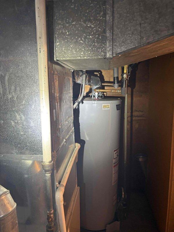 Completed inspection of residential water heater installation in basement mechanical room. Documented existing A.O. Smith water heater with standard plumbing connections, including brass fittings, valves, and both copper and black steel piping configurations. Photographed current system setup showing water heater positioned adjacent to heating system, with all connections, safety labels, and overhead ductwork visible for service records and future reference.