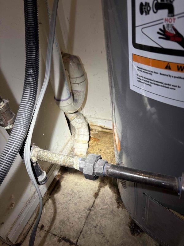 Responded to water leak investigation request in utility area near water heater where previous plumbers were unable to locate source. Conducted thorough inspection of water heater connections, supply lines, and surrounding plumbing infrastructure including kitchen sink waterline and refrigerator connection point. Upon examination, identified significant water damage and staining on concrete floor extending to wall junction, with visible moisture intrusion patterns and deterioration at baseboard level. Inspected white-painted supply piping with visible age-related discoloration and rust staining at floor penetrations. Documented condition of plumbing connections, pipe fittings, and appliance interfaces through photographic evidence showing extent of water damage in affected area. Assessed flexible supply lines, rigid piping, and all visible connections within utility space to determine leak origin point.