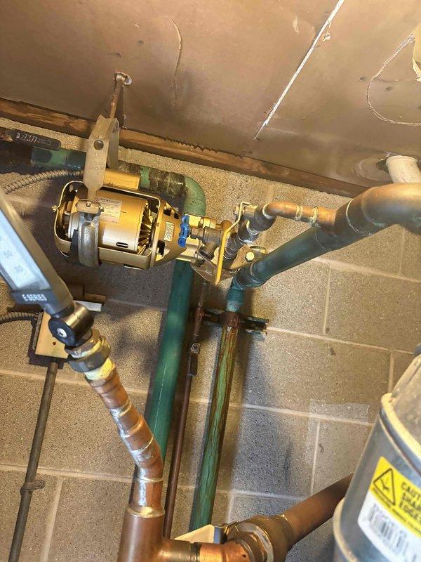 Responded to customer complaint of pounding noise from water heater. Upon inspection, identified an A.O. Smith residential water heater installed in utility closet with associated circulation pump system mounted overhead in adjacent basement area. The pounding noise was determined to be water hammer caused by pressure fluctuations in the plumbing system rather than an issue with the water heater unit itself. Examined the yellow electric circulator pump assembly with blue-handled isolation valves, copper supply lines, and older green-painted piping throughout the mechanical room. Verified water heater operation and proper venting through metallic flue pipe. Provided customer with estimate for installing water hammer arrestors on supply lines and recommended inspection of circulator pump mounting to reduce vibration transmission through the wooden beam support structure.