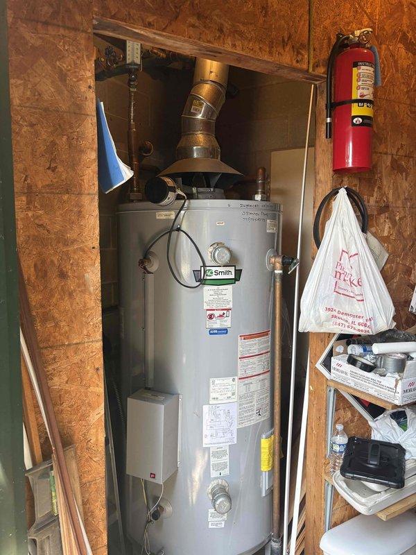 Responded to customer complaint of pounding noise from water heater. Upon inspection, identified an A.O. Smith residential water heater installed in utility closet with associated circulation pump system mounted overhead in adjacent basement area. The pounding noise was determined to be water hammer caused by pressure fluctuations in the plumbing system rather than an issue with the water heater unit itself. Examined the yellow electric circulator pump assembly with blue-handled isolation valves, copper supply lines, and older green-painted piping throughout the mechanical room. Verified water heater operation and proper venting through metallic flue pipe. Provided customer with estimate for installing water hammer arrestors on supply lines and recommended inspection of circulator pump mounting to reduce vibration transmission through the wooden beam support structure.