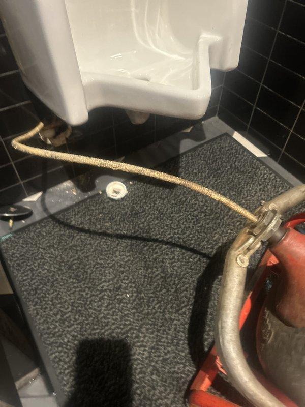 Responded to a clogged urinal fixture. Upon arrival, inspected the drainage system and identified blockage in the urinal waste line. Utilized drain cleaning equipment to clear the obstruction and restore proper flow. Verified fixture drainage functionality and confirmed complete resolution of the clog. Documented existing conditions including floor drain with mineral deposits and wear around plumbing connections.