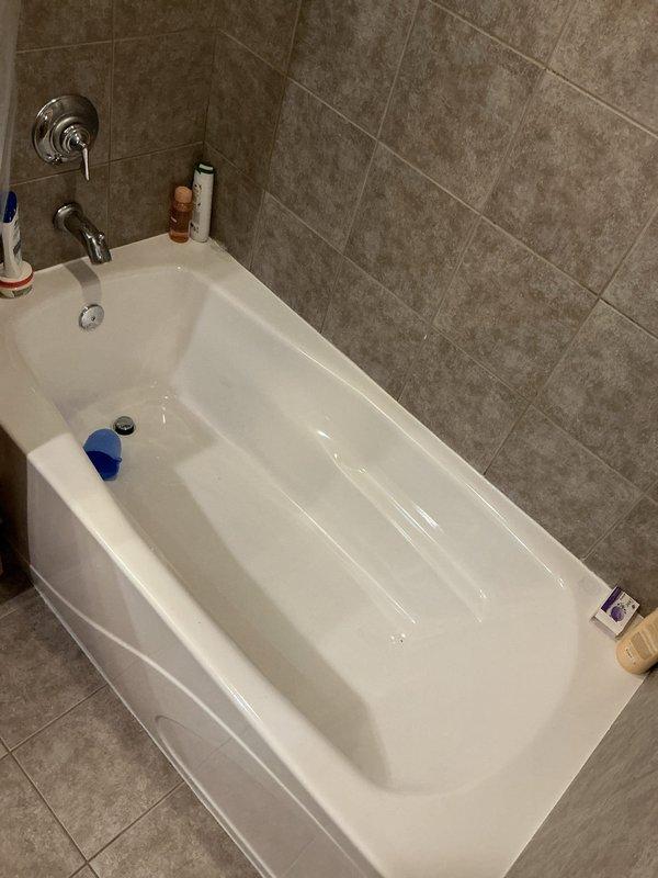 Responded to a water leak investigation in a high-rise residential unit where water was penetrating to the unit below. Upon arrival, inspected the bathroom tub area which had previously undergone drain replacement and subsequent resealing attempts. Conducted thorough visual inspection of the bathtub installation, surrounding tile work, and plumbing fixtures including chrome controls and tub spout. Examined the tub perimeter, grout lines, and caulking joints for potential breach points. Accessed the unit below to inspect ceiling penetration and plumbing infrastructure, documenting water damage patterns on concrete surfaces and identifying affected piping systems including insulated lines and copper supply lines. Photographed the bathtub installation showing corner alcove configuration with tile surround and documented the mechanical chase area below revealing pipe penetrations through ceiling structure. Investigation revealed ongoing water intrusion despite previous resealing work, indi