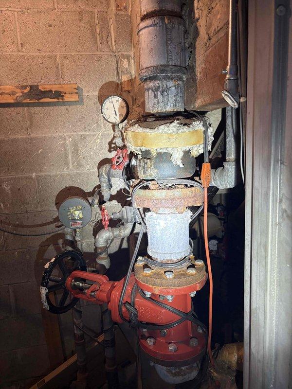 Coordinated building water shut down to facilitate plumbing system inspection and assessment. Documented existing conditions of main water supply assembly, including backflow preventer, pressure gauge, and associated piping showing deterioration of insulation wrapping and age-related wear. Inspected exposed garage ceiling area to assess horizontal piping system and identified significant water damage and potential mold growth on overhead plywood surfaces. Photographed before conditions of plumbing infrastructure and water-damaged ceiling areas for documentation and estimation purposes. Building management opened ceiling access to allow for comprehensive evaluation of garage horizontal piping network requiring potential replacement or repair work.