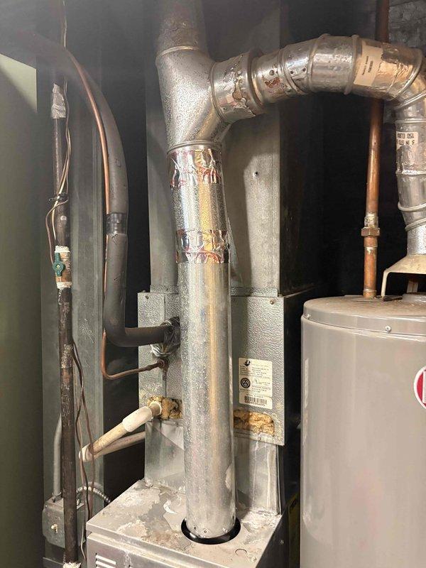 Responded to service call for water heater leaking from bottom. Upon inspection, found Rheem tanked water heater with visible signs of tank failure and bottom leakage. Documented existing installation configuration including connections to copper supply lines, gas line, temperature and pressure relief valve, and drainage system. Photographed equipment condition and surrounding utility area showing water heater positioned adjacent to furnace unit with associated ductwork and plumbing infrastructure. Advised customer that bottom leaks typically indicate internal tank corrosion and require full water heater replacement, as tank integrity cannot be restored once compromised.