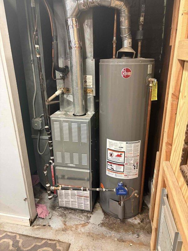 Responded to service call for water heater leaking from bottom. Upon inspection, found Rheem tanked water heater with visible signs of tank failure and bottom leakage. Documented existing installation configuration including connections to copper supply lines, gas line, temperature and pressure relief valve, and drainage system. Photographed equipment condition and surrounding utility area showing water heater positioned adjacent to furnace unit with associated ductwork and plumbing infrastructure. Advised customer that bottom leaks typically indicate internal tank corrosion and require full water heater replacement, as tank integrity cannot be restored once compromised.
