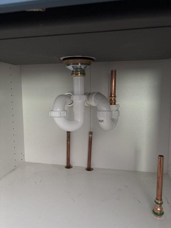 Completed repipe of lower kitchen drain assembly beneath sink. Accessed plumbing system through cabinet cutout to install new white PVC P-trap configuration with chrome strainer basket and vertical tailpiece. Installation included copper supply line connections with green and brass shut-off valves. All drain components properly fitted to undermount stainless steel sink basin in white marble-style countertop. System tested and verified operational with no leaks detected.