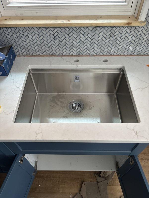 Completed repipe of lower kitchen drain assembly beneath sink. Accessed plumbing system through cabinet cutout to install new white PVC P-trap configuration with chrome strainer basket and vertical tailpiece. Installation included copper supply line connections with green and brass shut-off valves. All drain components properly fitted to undermount stainless steel sink basin in white marble-style countertop. System tested and verified operational with no leaks detected.