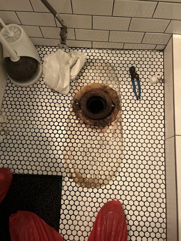 Responded to water leak complaint from unit below affecting bathroom ceiling fan. Upon inspection, discovered heavily corroded and rusted toilet flange in second-floor bathroom directly above affected area. The deteriorated flange showed significant rust damage extending onto surrounding hexagonal tile floor, confirming the source of water infiltration. Removed toilet to access and assess flange condition, documenting extensive corrosion that compromised the seal between toilet and drain pipe, allowing water to leak through to the lower unit's ceiling during toilet use.