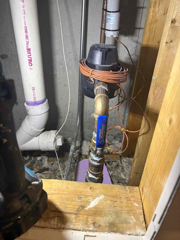 Installed complete plumbing assembly in utility area including PVC DWV piping system with vertical configuration and elbow joints. Mounted pump unit with coiled copper tubing and integrated blue-labeled pressure regulator component on wooden platform base. Installed brass pipe assembly with ball valve for shut-off control and secured electrical wiring along installation path. All connections properly fitted and assembly positioned for optimal access and serviceability in mechanical space.