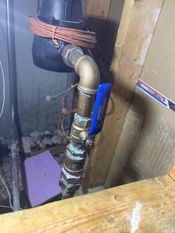 Installed complete plumbing assembly in utility area including PVC DWV piping system with vertical configuration and elbow joints. Mounted pump unit with coiled copper tubing and integrated blue-labeled pressure regulator component on wooden platform base. Installed brass pipe assembly with ball valve for shut-off control and secured electrical wiring along installation path. All connections properly fitted and assembly positioned for optimal access and serviceability in mechanical space.