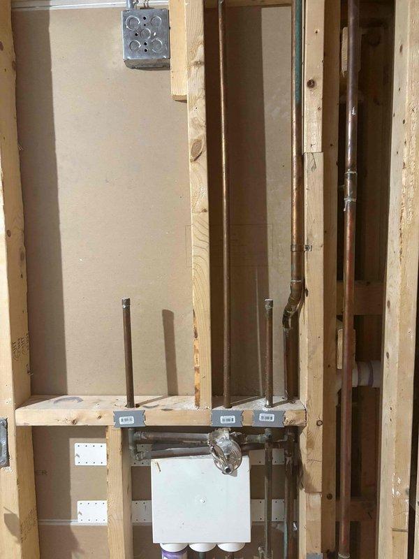 Completed plumbing rough-in installation in an interior renovation area featuring exposed brick wall and new wood framing. Installed white PVC drainage piping system with multiple horizontal and vertical runs, including elbow fittings secured to wooden studs, and staged copper water supply risers with mounting brackets on a horizontal board for future fixture connections.